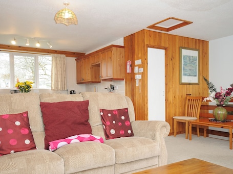 A cozy living room with a beige sofa, wooden walls, a dining area with a wooden table and chairs, and a kitchen with wooden cabinets.