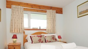 Een netjes ingerichte slaapkamer met een houten hoofdbord, een raam met bloemen gordijnen, een nachtkastje met een lamp, en een bed met bloemen kussens en handdoeken.