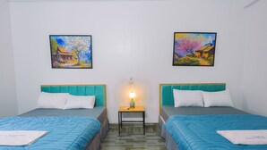 Quarto de hotel com duas camas, cada uma com cabeceira azul-turquesa e roupa de cama branca. Há duas pinturas nas paredes, uma representando uma casa com uma árvore e a outra uma paisagem de vila. Uma pequena mesa com uma luminária fica entre as camas.