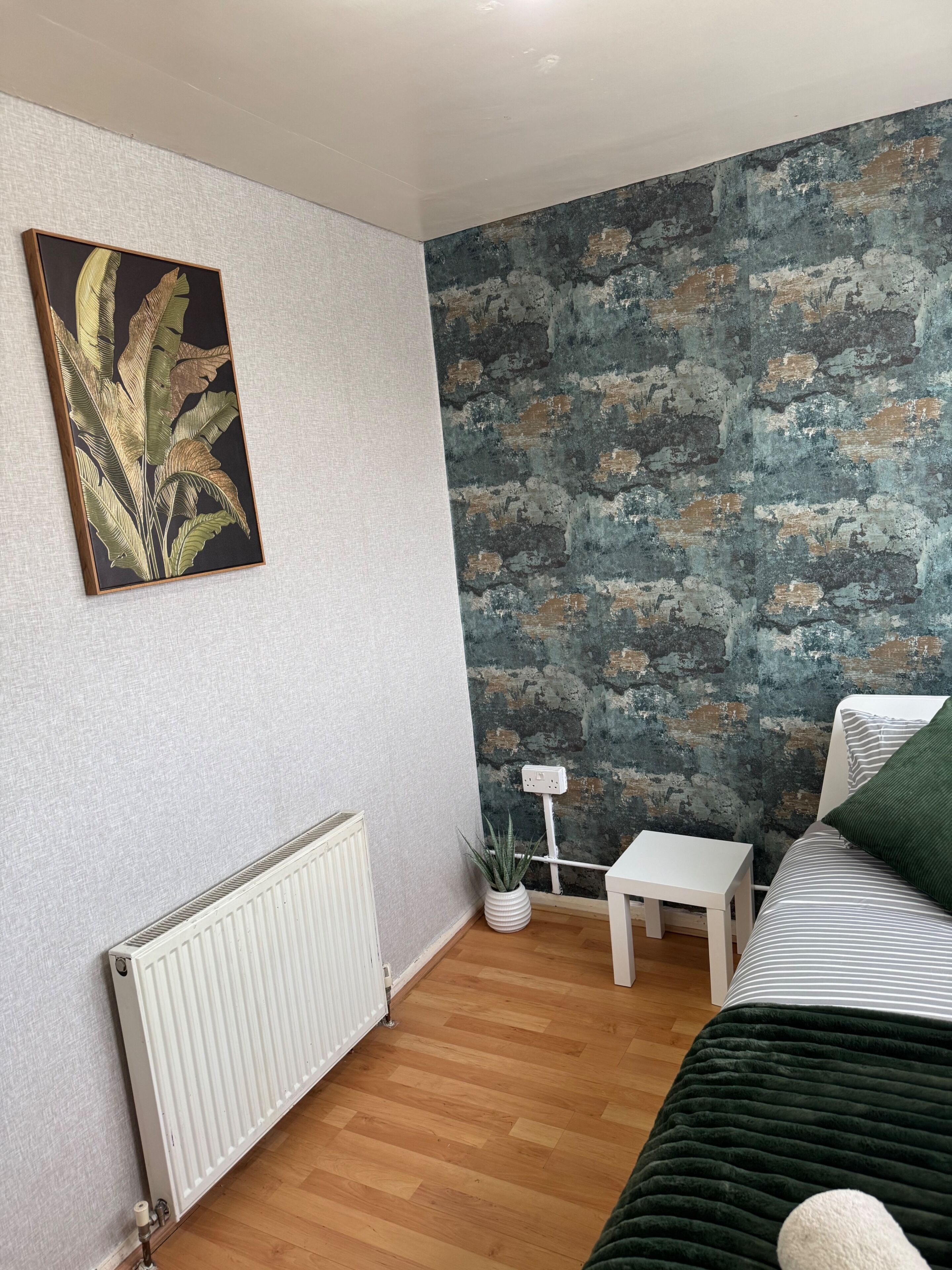 Ein Zimmer mit Heizkörper, einem kleinen weißen Tisch, einer Topfpflanze, einem Bett mit einem gestreiften Kissen und einer Wand mit strukturierter Oberfläche.
