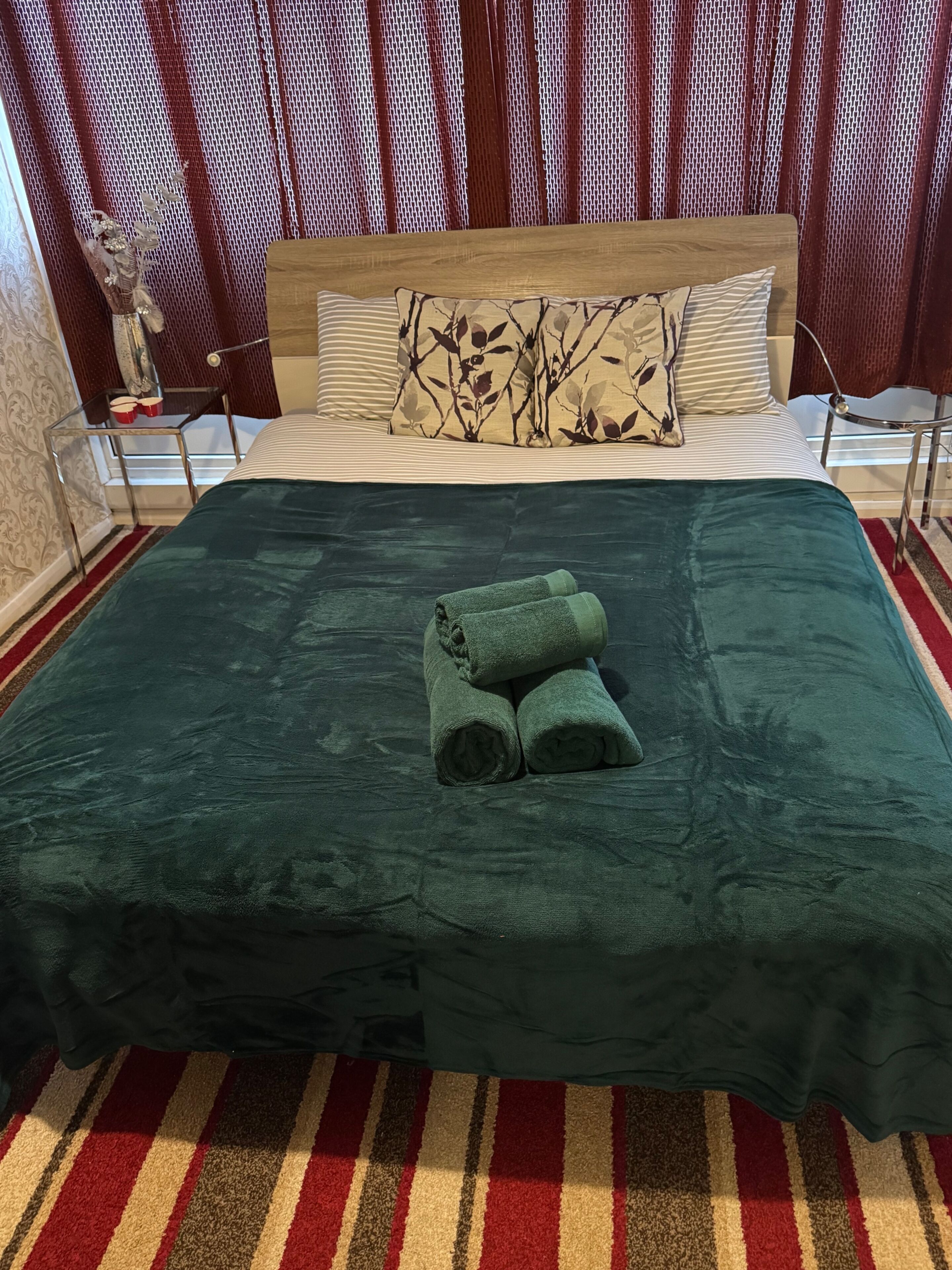 A bed with a dark green bedspread, two rolled towels, and a striped pillow.