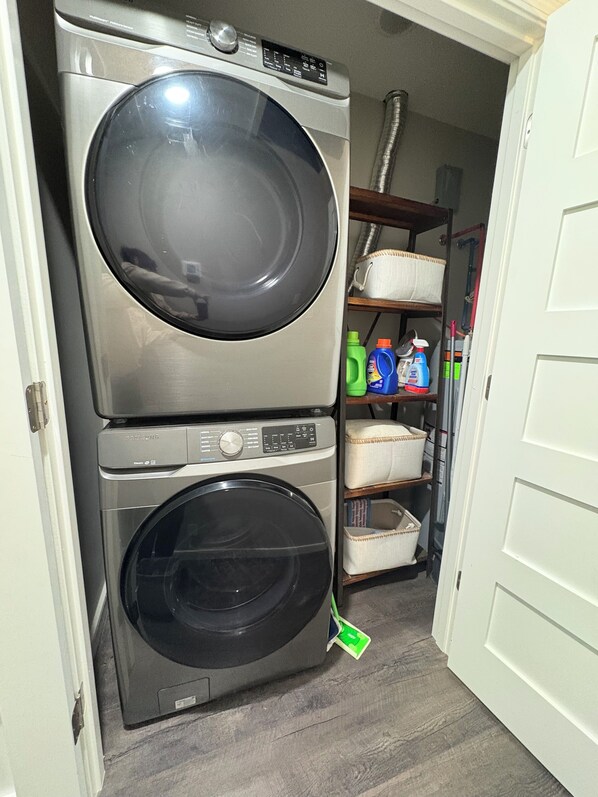 A laundry area with stacked washers and dryers, shelves with cleaning supplies, and a door leading to another room.