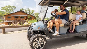 A golf cart with a driver and passengers on a paved road, with a wooden building and trees in the background.