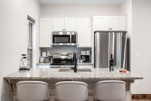 A modern kitchen with white cabinetry, stainless steel appliances, a granite countertop, and three bar-style chairs.