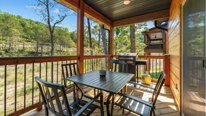A covered outdoor deck with a table and chairs, a grill, and a view of the surrounding forest.