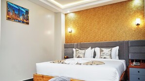 A modern hotel room with a wooden headboard, a large bed with white linens, a grey bench, and a cityscape painting on the wall.