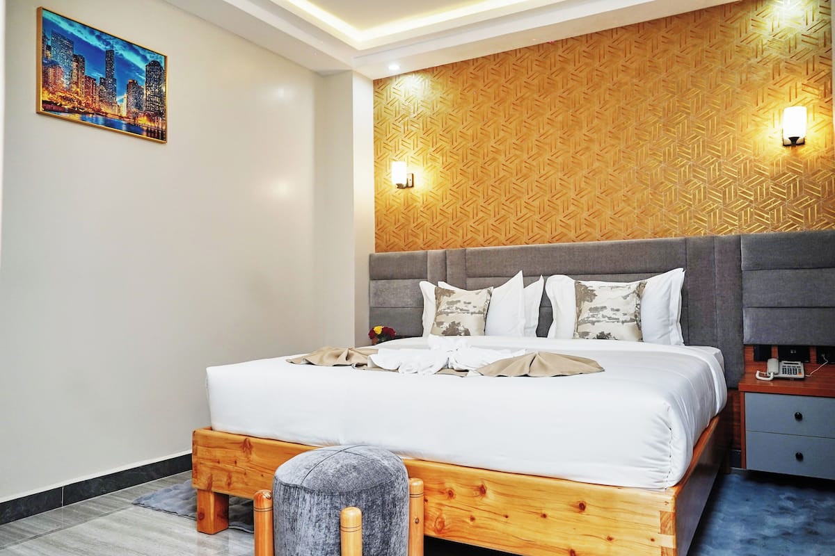 A modern hotel room with a wooden headboard, a large bed with white linens, a grey bench, and a cityscape painting on the wall.