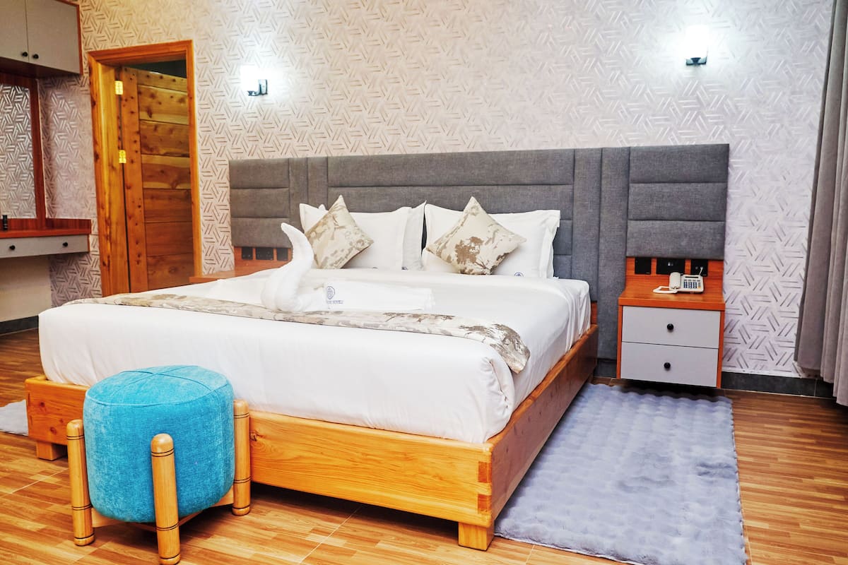 A neatly made bed with a grey headboard, a wooden nightstand with a lamp, and a blue ottoman.