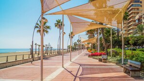 Een boulevard aan zee met beschaduwde zitplekken, palmbomen en een strand op de achtergrond.