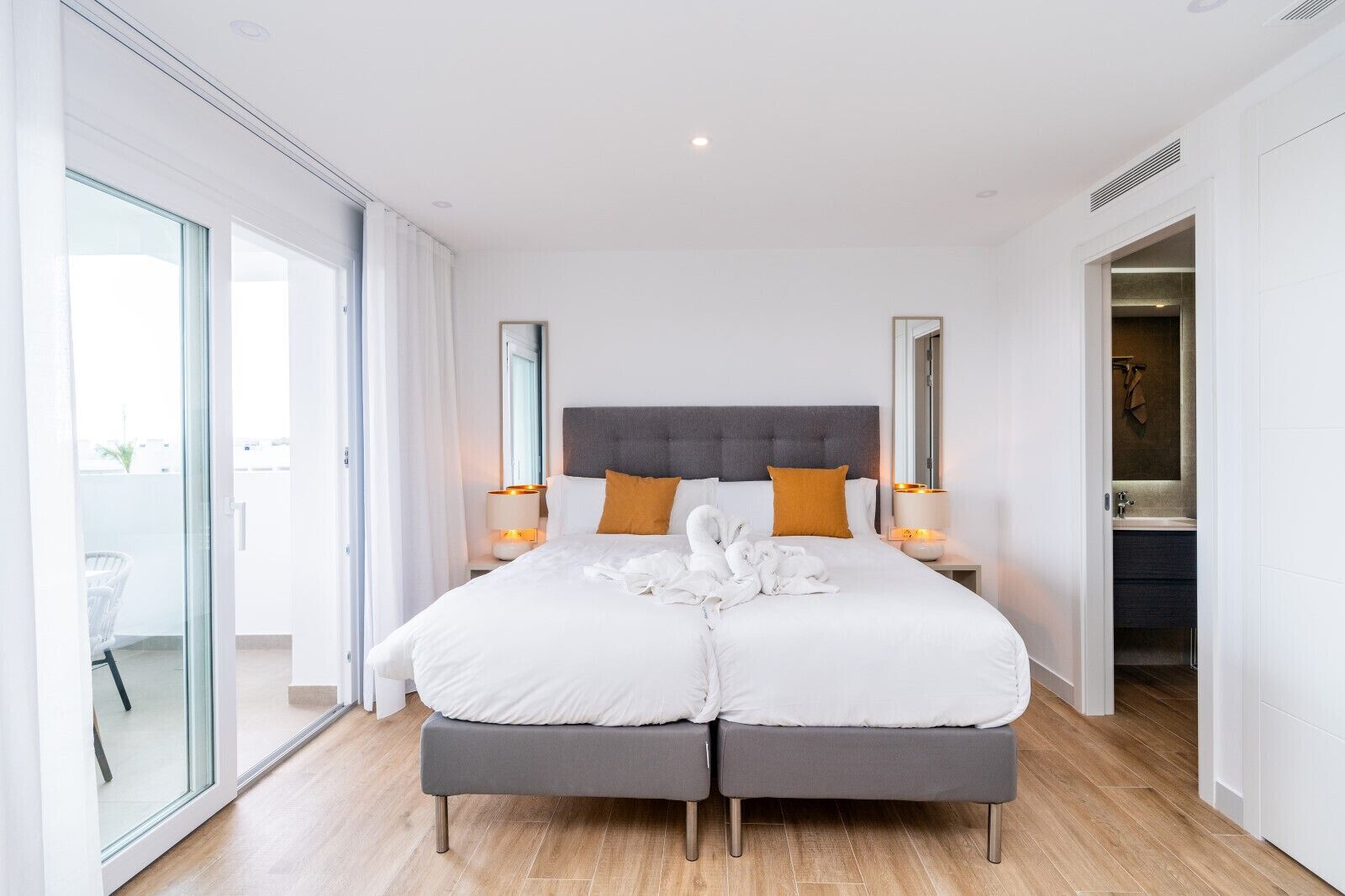 Quarto de hotel moderno com uma cama grande, duas mesas de cabeceira com luminárias, um espelho grande e uma porta de vidro deslizante que dá para uma varanda.