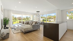 Un salon moderne avec un canapé gris, une table basse et un téléviseur. Une grande fenêtre offre une vue sur un jardin et un balcon.