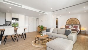 Ein modernes Wohnzimmer mit einer Couch, einem Hocker und einem Couchtisch, ein Essbereich mit Stühlen und Tisch sowie eine Küche mit Geräten.