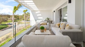 Ein Balkon mit einer weißen Couch, einem kleinen Tisch mit einer Pflanze und Blick auf eine Straße und Grünflächen.