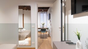 A modern hotel room with a wooden floor, a bed with white linens, a desk with a vase of flowers, and a TV mounted on the wall.