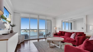 A spacious living room with a large window, a red sofa, a round coffee table, and a view of the sea. - Le Petit Italie Water View 2 2 With Pool (Sunny Isles Beach)