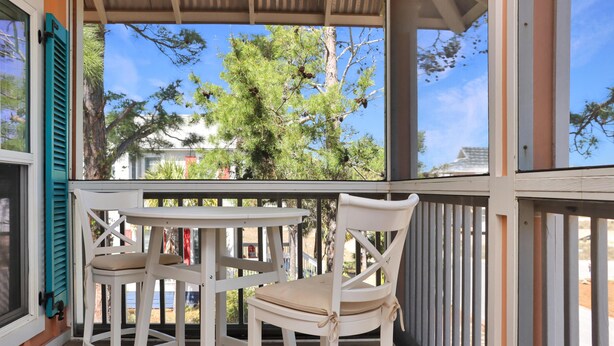 A covered outdoor seating area with a white table and chairs, overlooking a tree and a building.