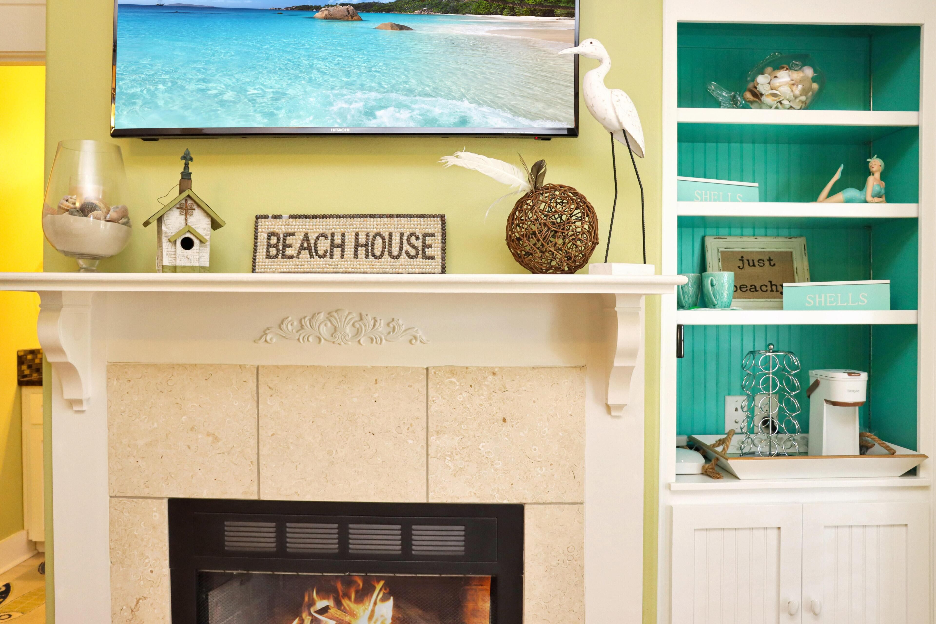 Ein Kamin mit einem 'Beach House' Schild, darüber ein Fernseher und eine Ablage mit Dekoration.