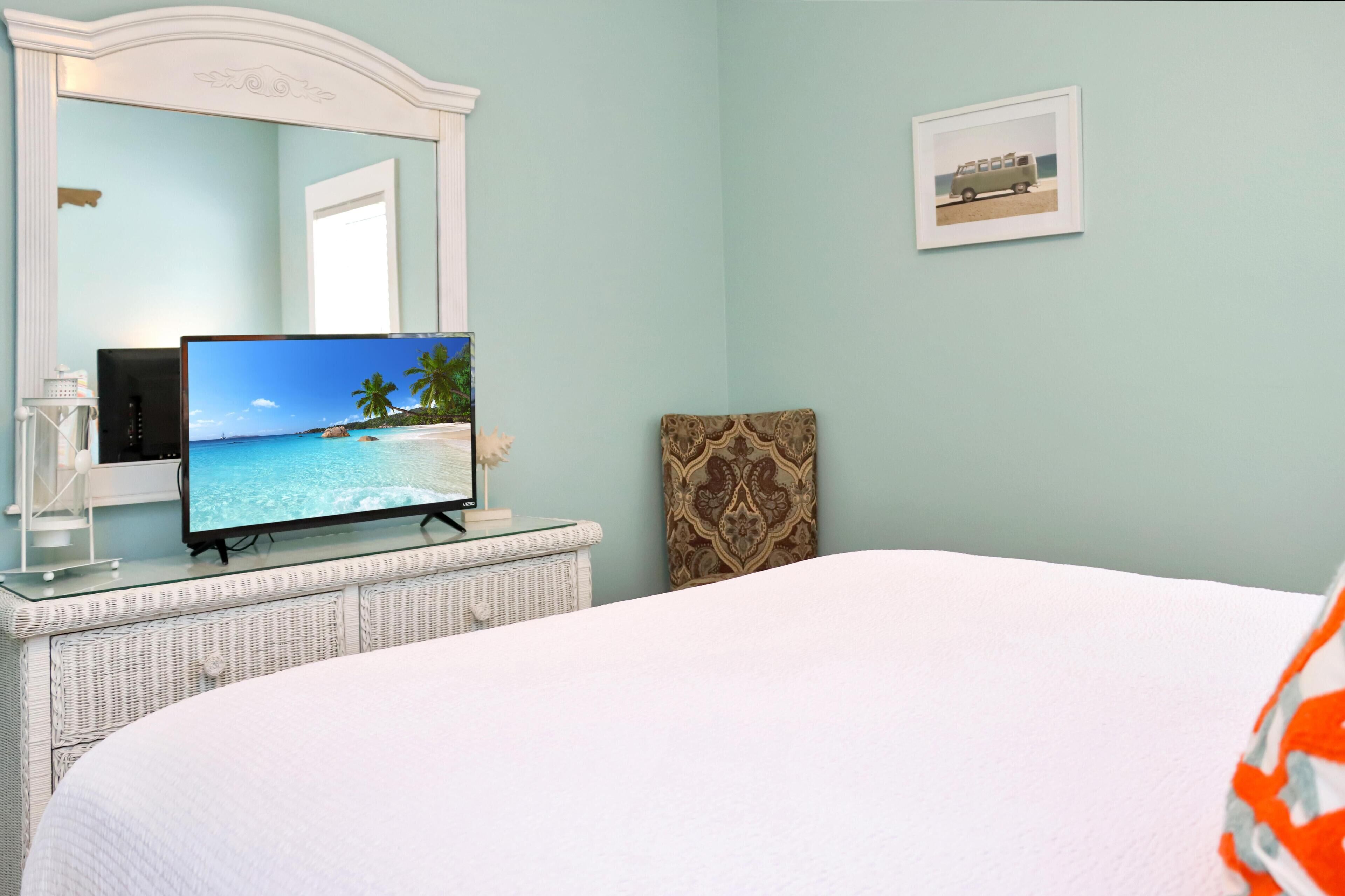 A bedroom with a bed, a television on a glass-top dresser, a mirror, and a framed picture on the wall.