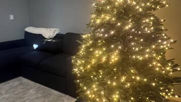 Un sapin de Noël décoré de guirlandes lumineuses et orné d’un nœud rouge, placé sur un tapis dans une pièce où se trouvent un canapé de couleur sombre et un luminaire au plafond.