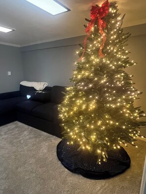 En julgran med ljus och ett rött band, placerad på ett golv med matta i ett rum med en mörkfärgad soffa och en taklampa.
