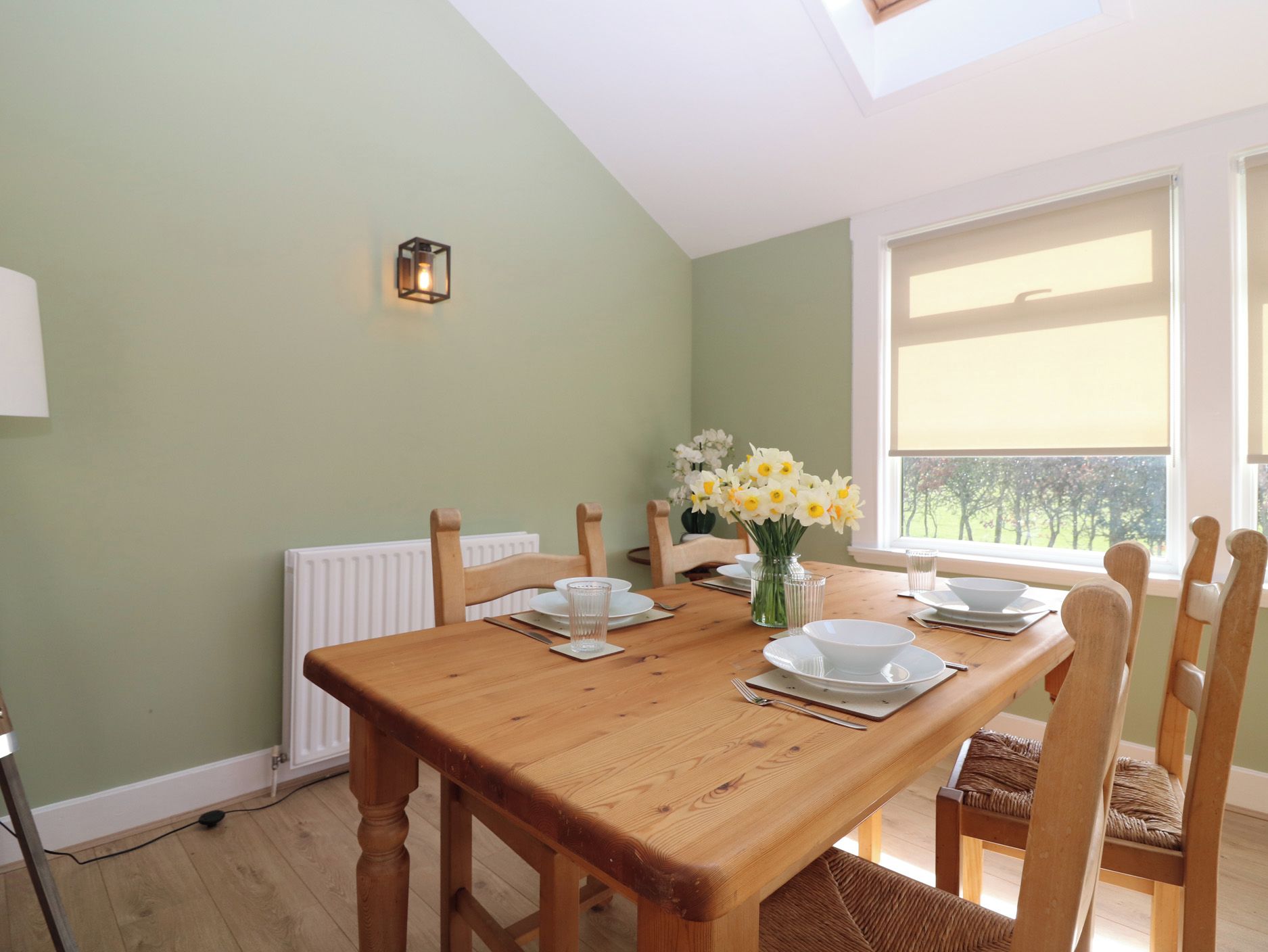 A dining area with a wooden table set for four, a vase of flowers, and a view of the outdoors through a window.