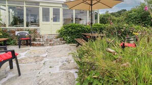 A stone patio with outdoor seating, a conservatory with glass doors, and a garden area with various plants and flowers. - Nanvivian (Truro)