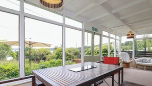 A covered outdoor seating area with a wooden table and chairs, large windows, and a view of greenery and a body of water. - Nanvivian (Truro)