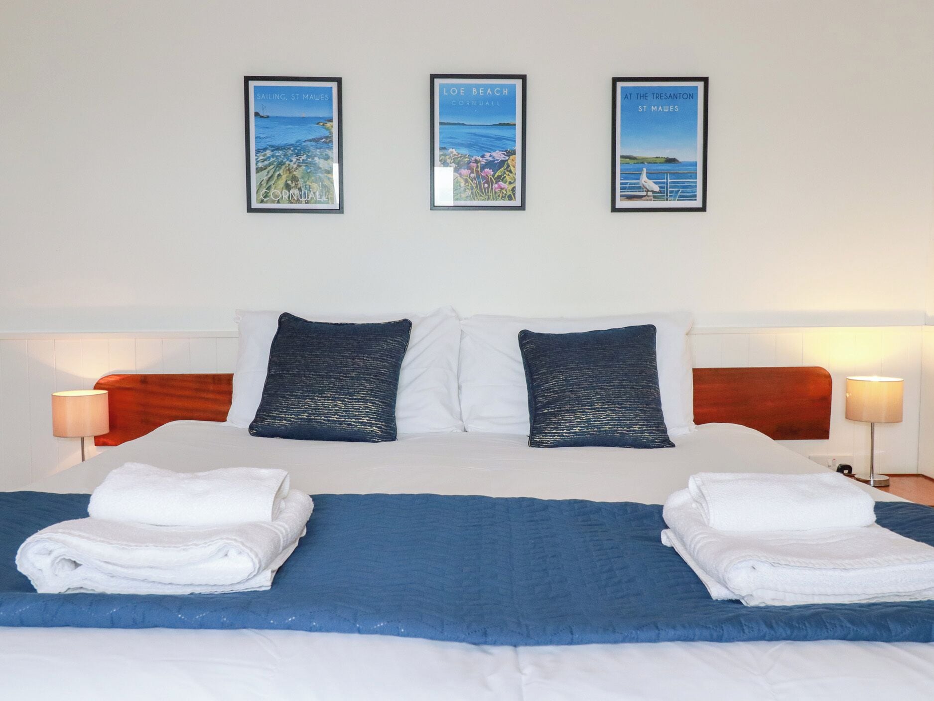 A hotel room with a bed, two pillows, a blue bedspread, two white towels, and three framed pictures on the wall.