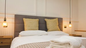 A neatly made bed with a checkered bedspread, two pillows, and a stack of folded towels on a bedside table.