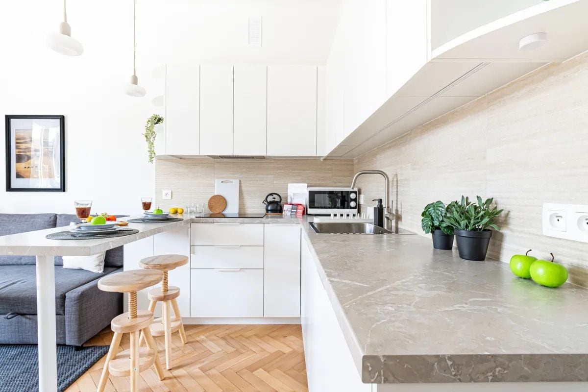 Une cuisine moderne avec des meubles blancs, un plan de travail en marbre et un coin repas comprenant un canapé gris et des tabourets en bois.