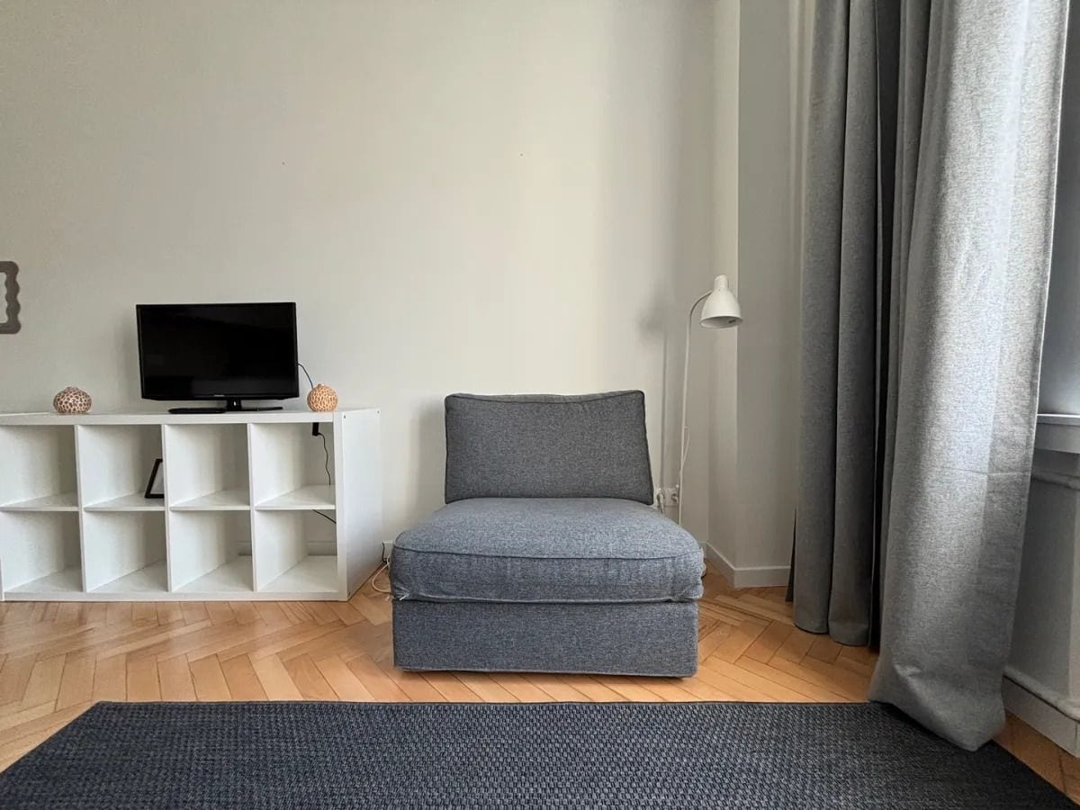 Una sala de estar con un sofá gris, un televisor sobre un estante blanco y una lámpara.