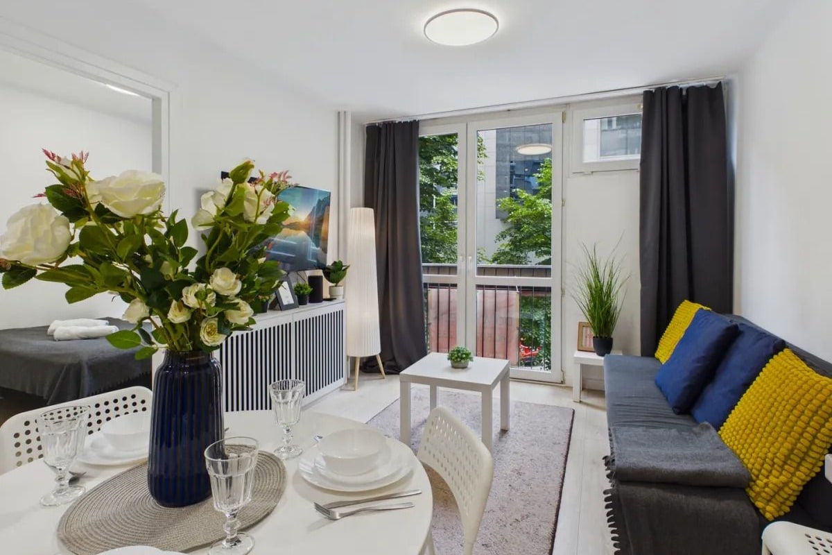 En moderne stue med spiseplads, en sofa med gule puder og et stort vindue med gardiner.