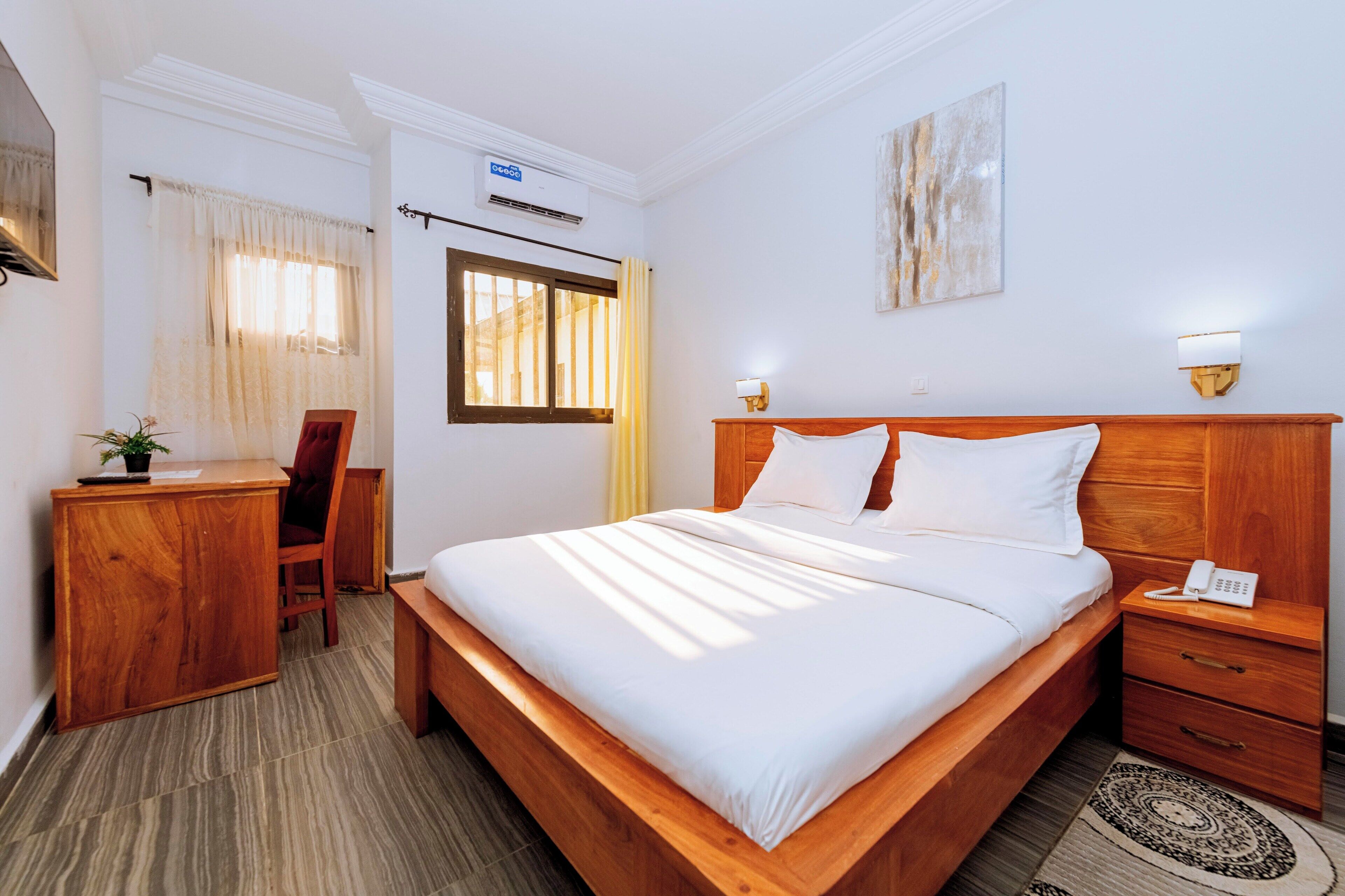 A hotel room with a wooden bed, a desk, a chair, a bedside table with a phone, and a wall-mounted lamp.