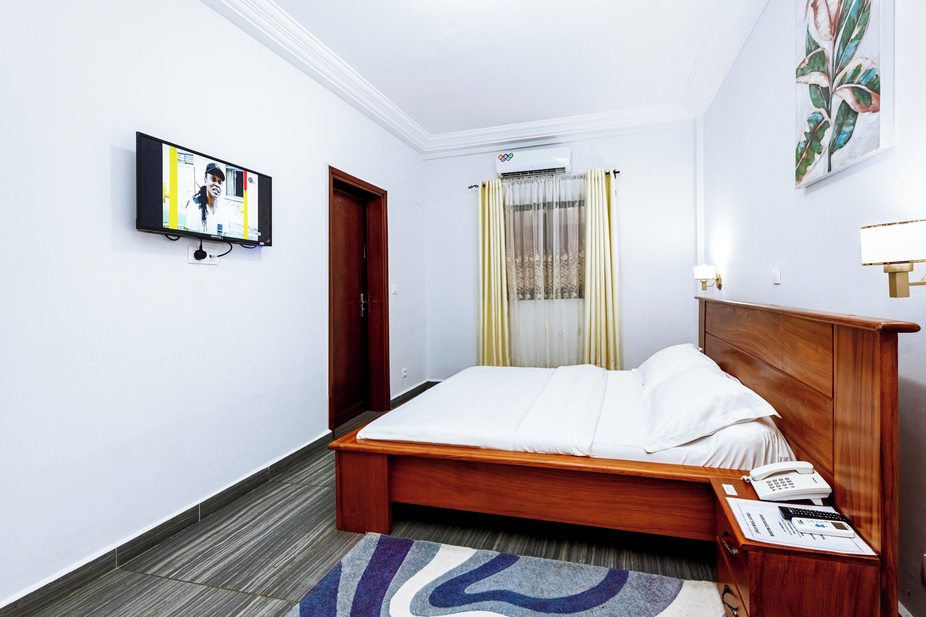 A hotel room with a wooden bed, a flat-screen TV mounted on the wall, a bedside table with a phone and papers, and a window with curtains.
