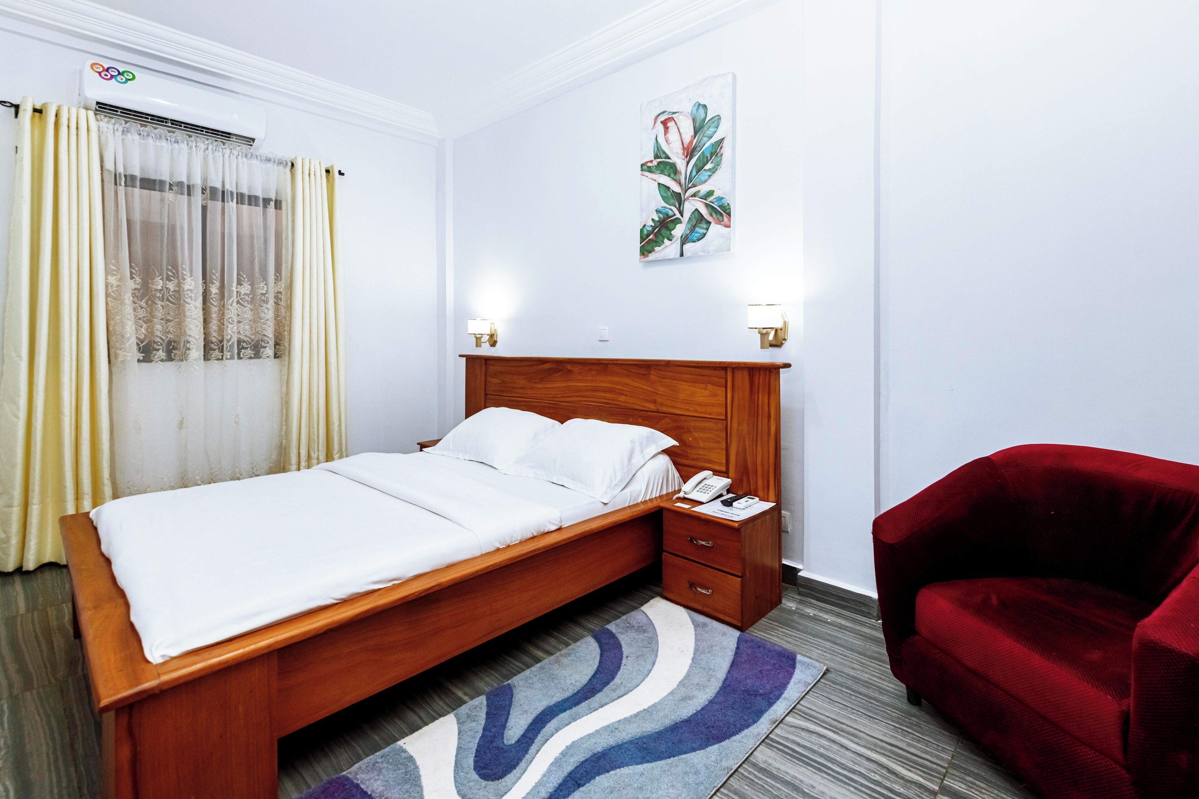 A hotel room with a wooden bed, a red armchair, a telephone, and a wall-mounted air conditioner.