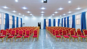 Ein Konferenzraum mit Reihen roter Stühle und blauen Vorhängen, einem Projektor an der Decke und einem gefliesten Boden.
