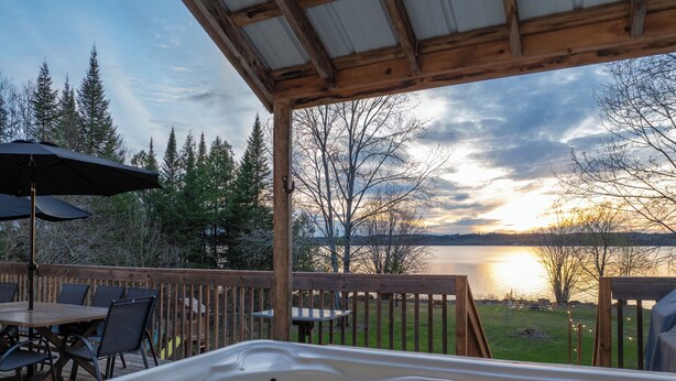 Ein Whirlpool mit Holzterrasse und Sitzgelegenheiten, mit Blick auf einen See, Bäume und einen Sonnenuntergang.
