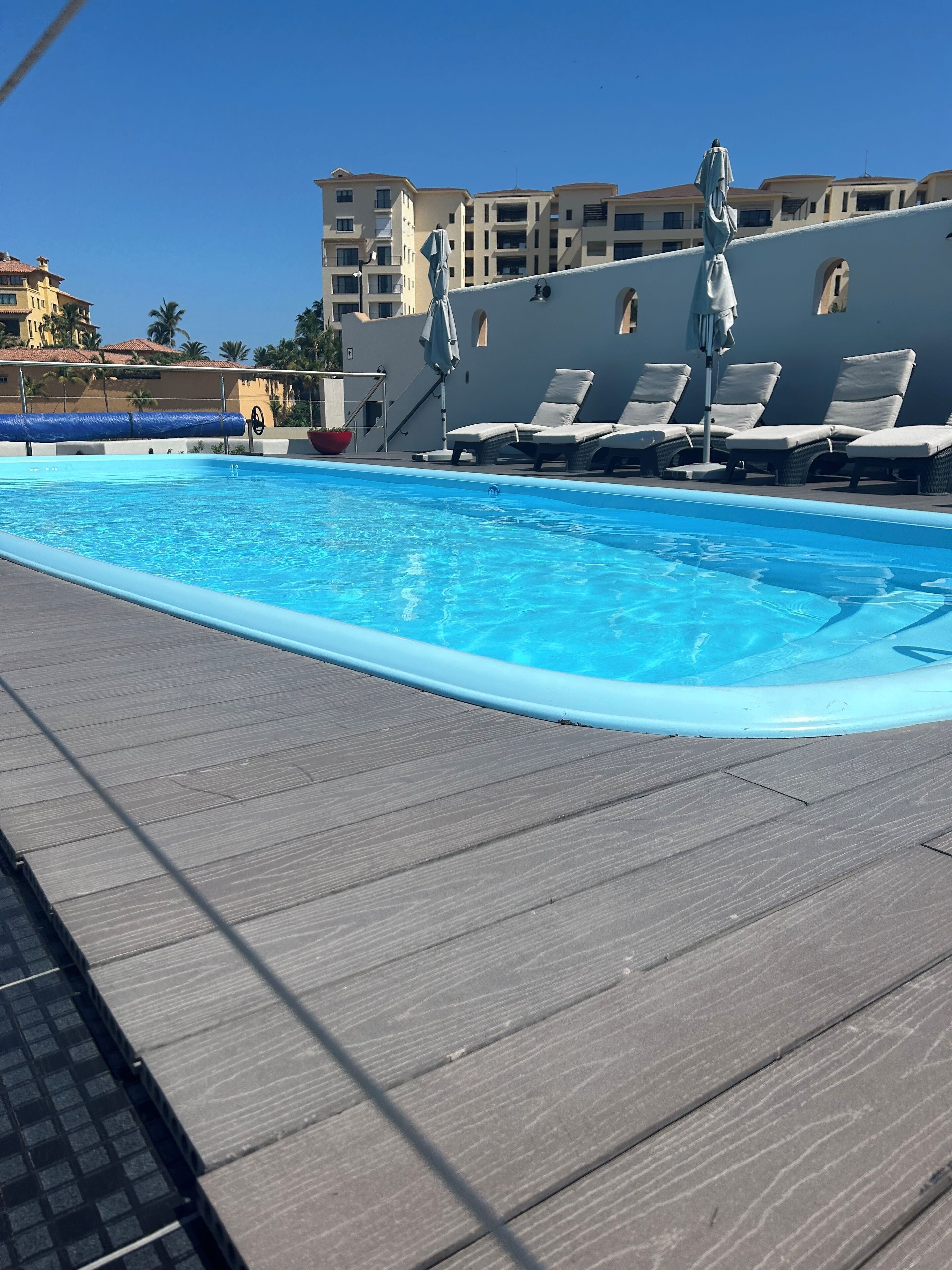 Une piscine entourée d’une terrasse en bois et d’un bâtiment doté de balcons, avec des chaises longues et des parasols à proximité.