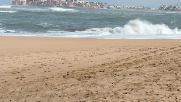 A beach with waves crashing onto the shore and a cityscape in the background.