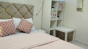 A bedroom with a bed, a nightstand, a shelf with decorative items, and a wall-mounted light fixture.