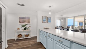 Una cucina moderna con armadietti azzurri, un piano di lavoro bianco e un pavimento in legno. Si vede una finestra con vista sulla spiaggia, una pianta sul bancone e un'area pranzo attraverso una porta aperta.