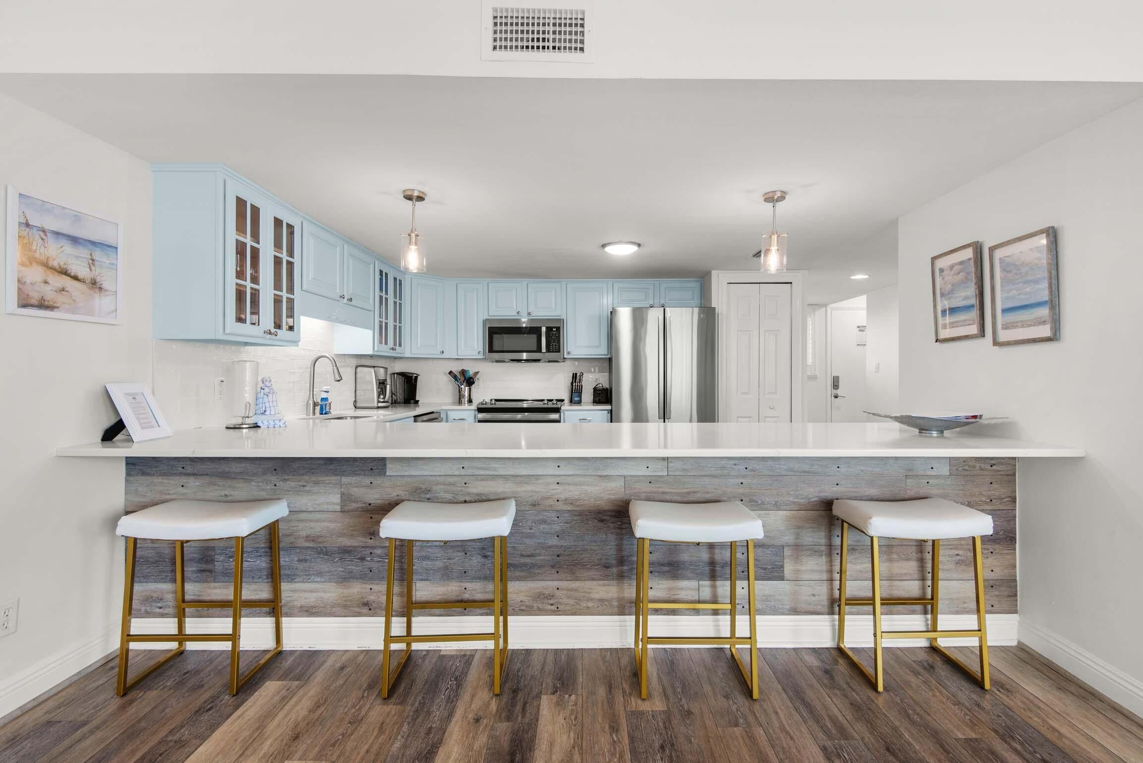 A modern kitchen with light blue cabinets, a white island with stools, stainless steel appliances, and a wooden floor.