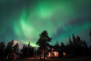 A cabin with a porch and a car parked in front, surrounded by trees under a night sky with the aurora borealis. - Camp Alta Kiruna (Kiruna)