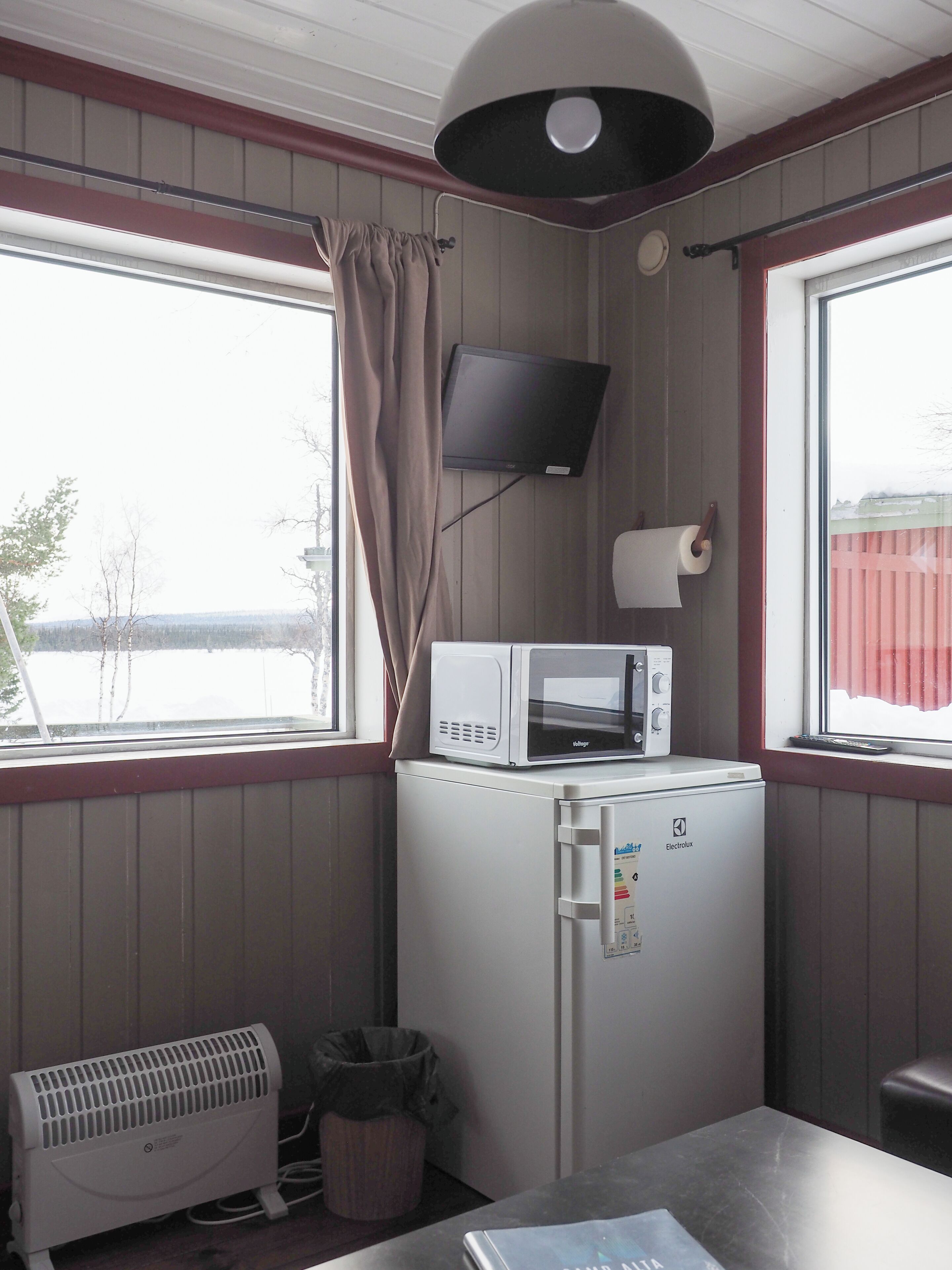 A compact kitchenette with a microwave, a small refrigerator, a heater, and a TV mounted on the wall.
