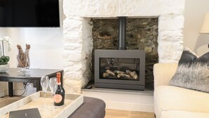 A living room with a stone fireplace, a flat-screen TV mounted on the wall, a sofa, and a coffee table with a bottle of wine and glasses. - 3 Rosemary Lane (Conwy)