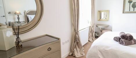 Une chambre avec un grand miroir rond, une table de chevet avec une lampe, un lit avec des serviettes pliées, une petite table de chevet et une fenêtre avec des rideaux.