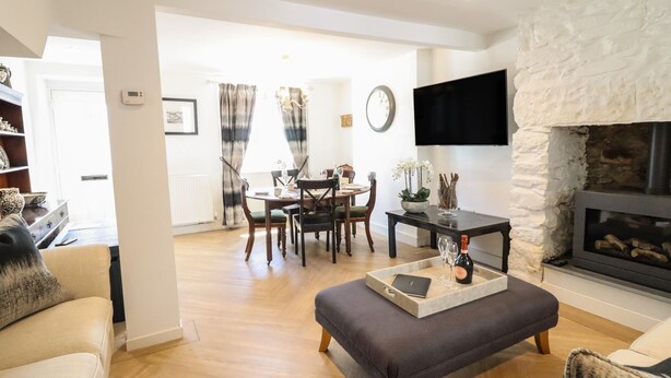 A modern living room with a fireplace, a dining area with a chandelier, and a flat-screen TV mounted on a stone wall. - 3 Rosemary Lane (Conwy)