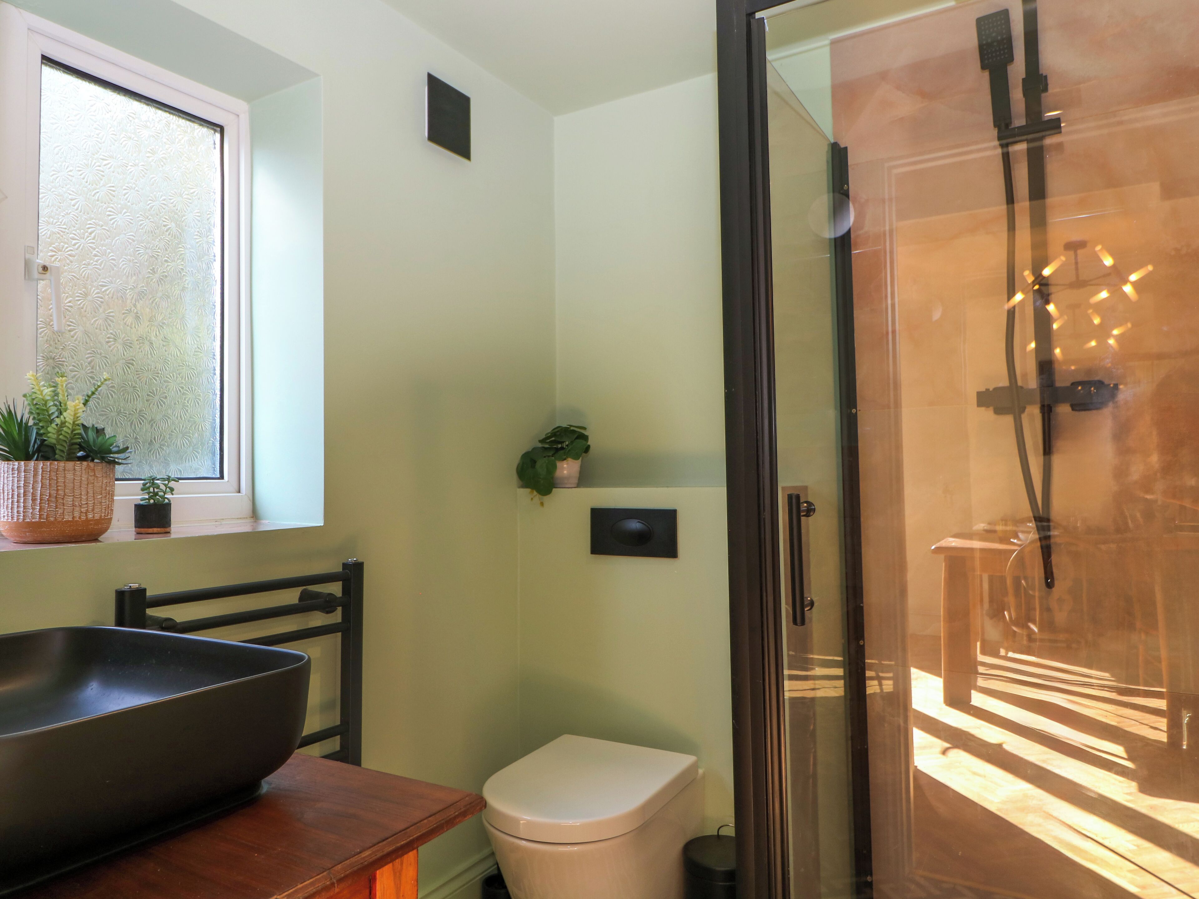 A modern bathroom with a glass shower enclosure, a toilet, a sink with a large basin, and two potted plants on the windowsill.