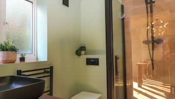 A modern bathroom with a glass shower enclosure, a toilet, a sink with a large basin, and two potted plants on the windowsill.