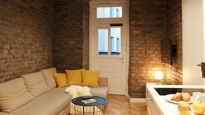 A cozy living room with a brick wall, a sofa with yellow cushions, a small round table with a book, and a door leading outside. - Cracow Fancy Brick #4 - Old Town (Kraków)